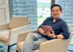 Desmond Yong, sitting on a chair and holding an iPad. He is wearing glasses, dark blue shirt, and jeans.